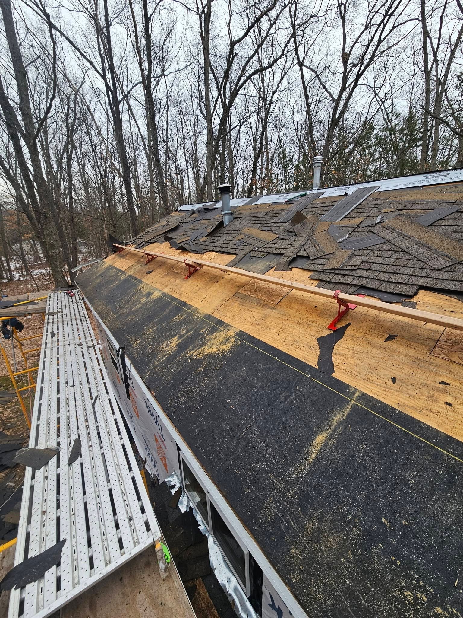 Partially stripped roof with exposed wood and new shingles, viewed beside leafless trees