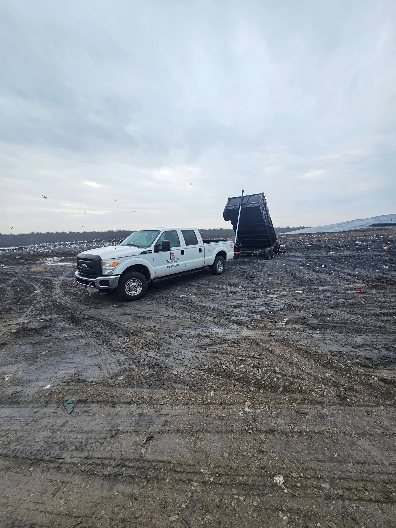 White pickup truck with a dump trailer on a muddy dirt site under a cloudy sky