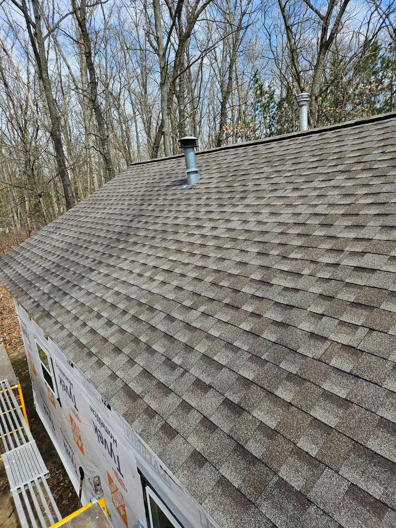 Weathered shingle roof with a vent pipe, viewed at an angle against leafless trees and blue sky