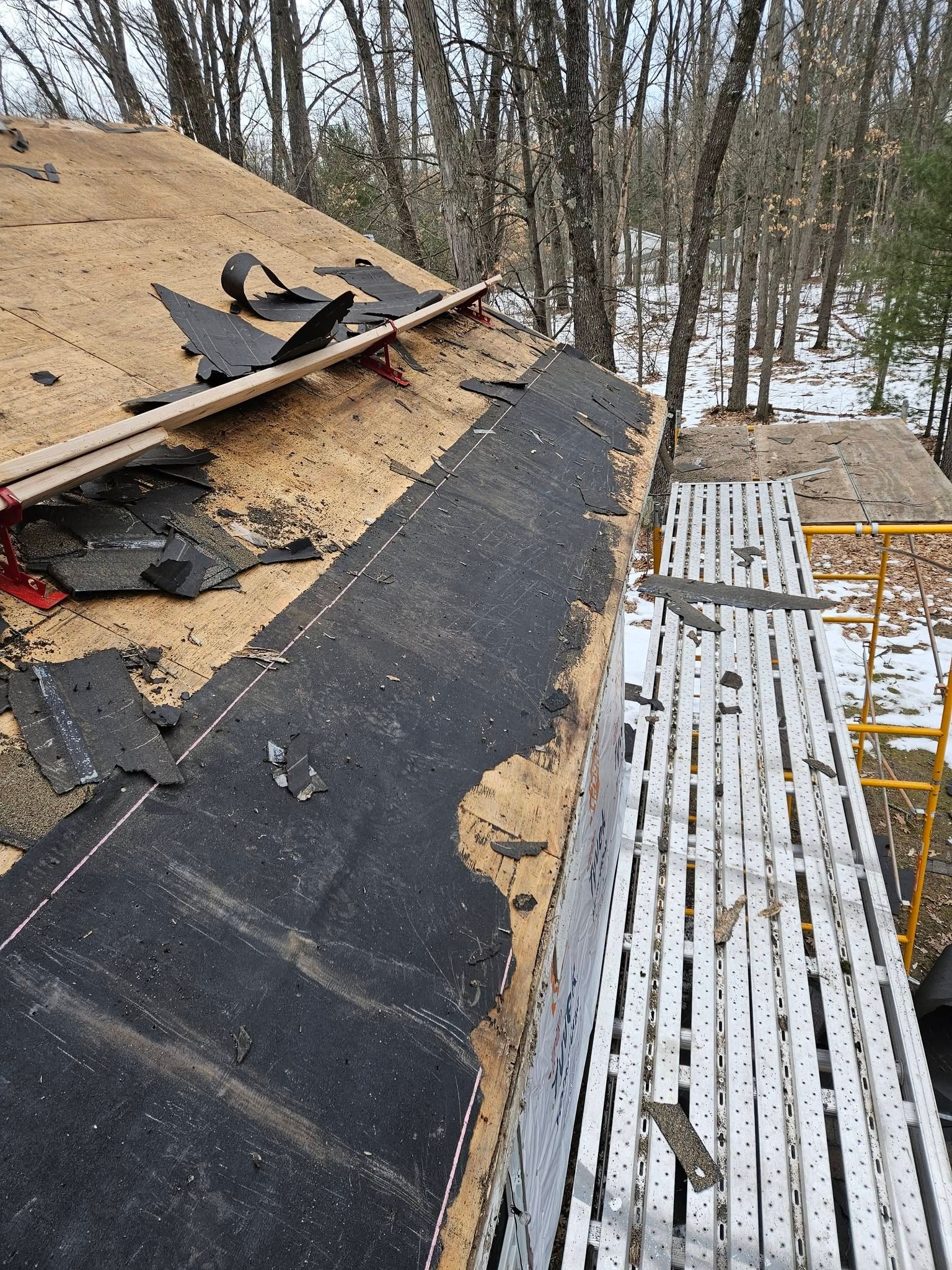 Roof under repair with black underlayment and scattered shingles beside a snowy wooded yard