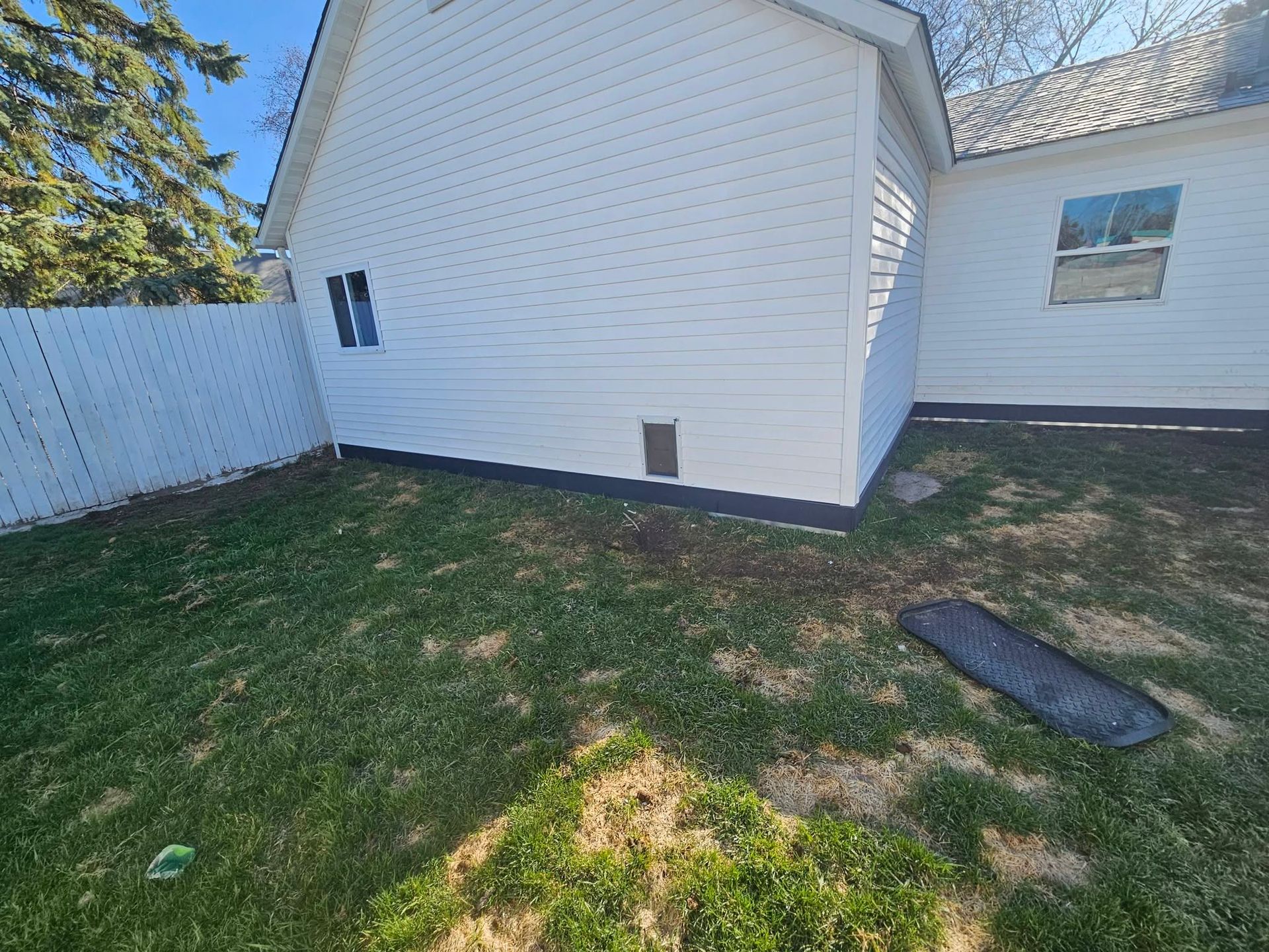 Backyard with white house siding, patchy grass, and a wooden fence on the left.