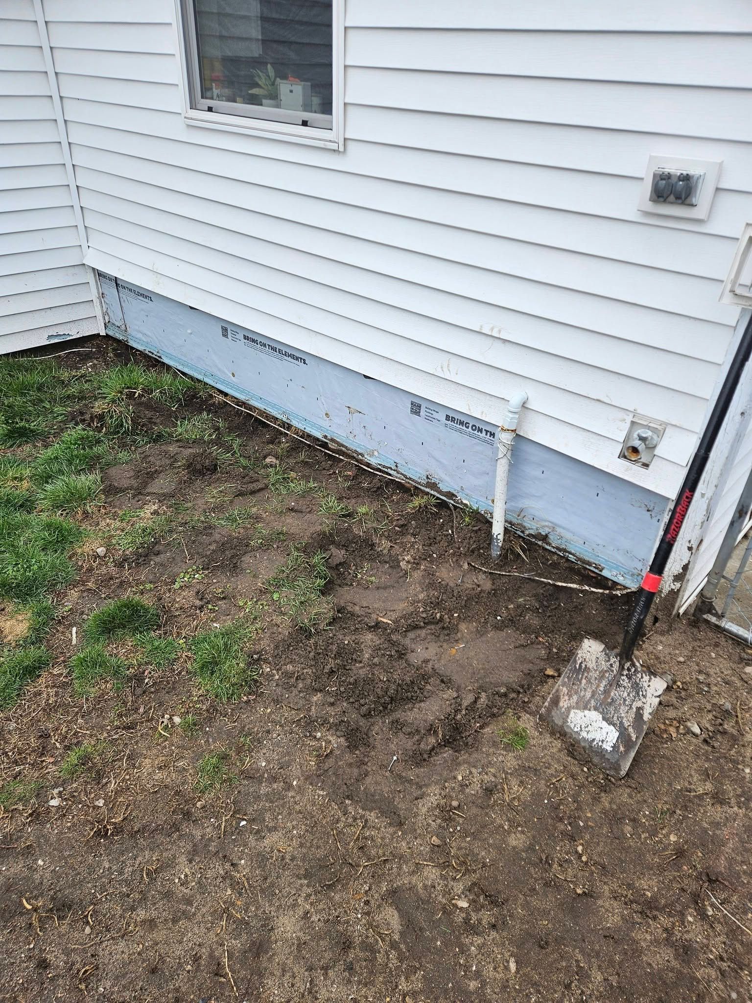 Bare dirt strip beside white house siding, with a shovel and utility fixtures near the foundation.