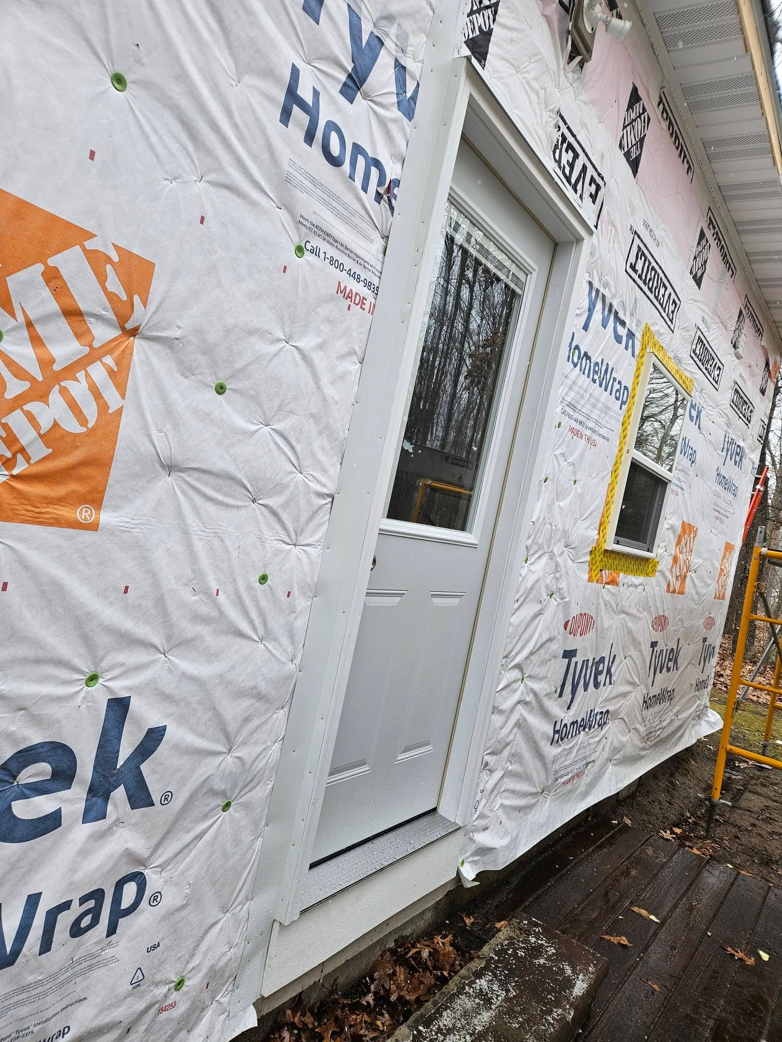 House wrapped in white Tyvek with a gray door and windows along a muddy walkway