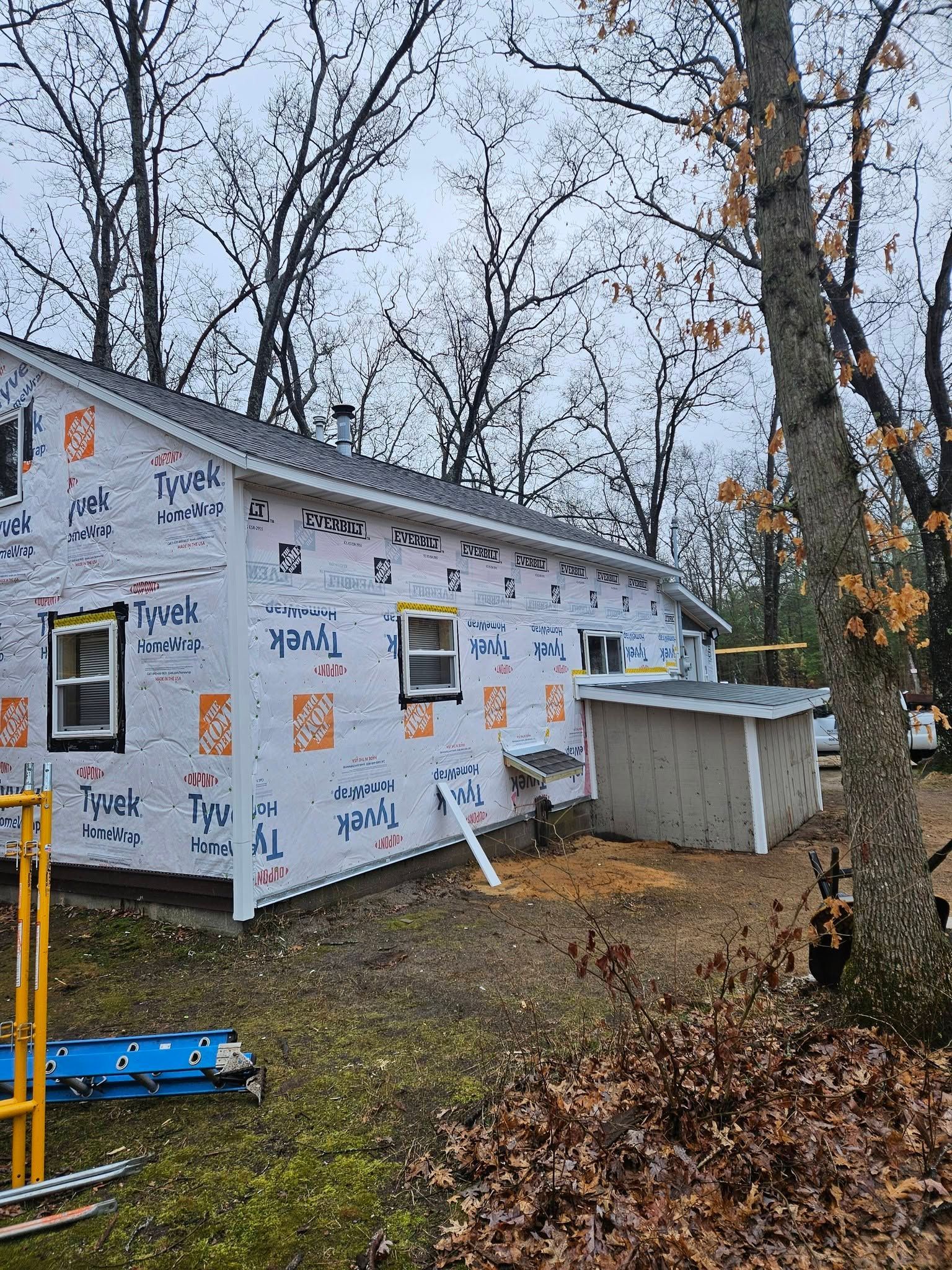 House under construction wrapped in white house wrap, with bare trees and a small shed on a grassy lot.