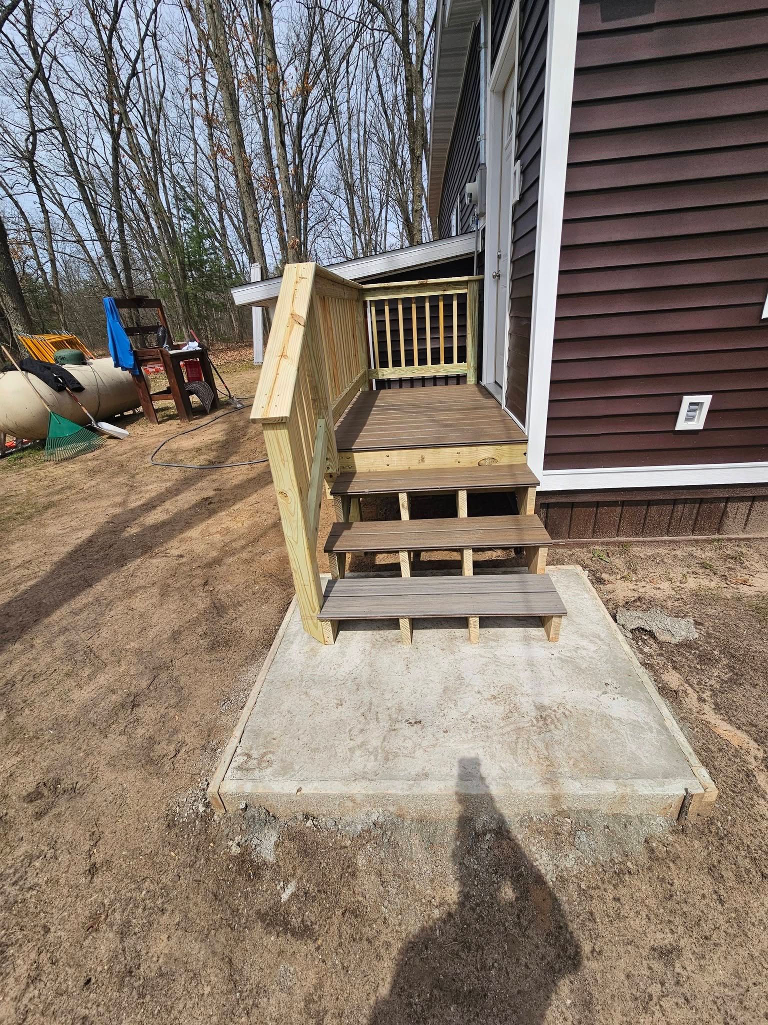 Wooden front steps with railing leading to a house entrance beside a concrete pad and bare yard.
