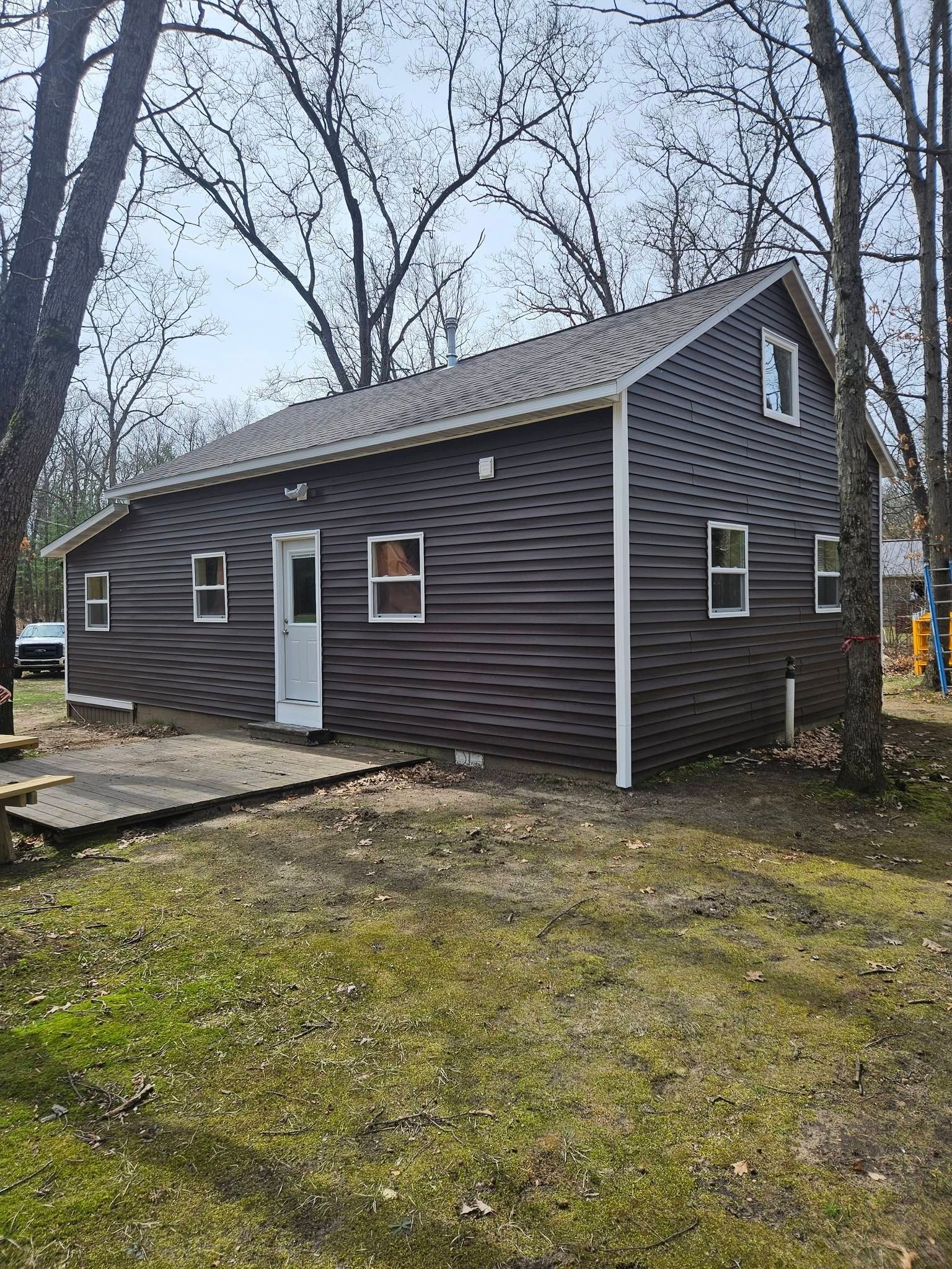 Small dark-sided house with white trim, front door, windows, and a leafless yard on an overcast day