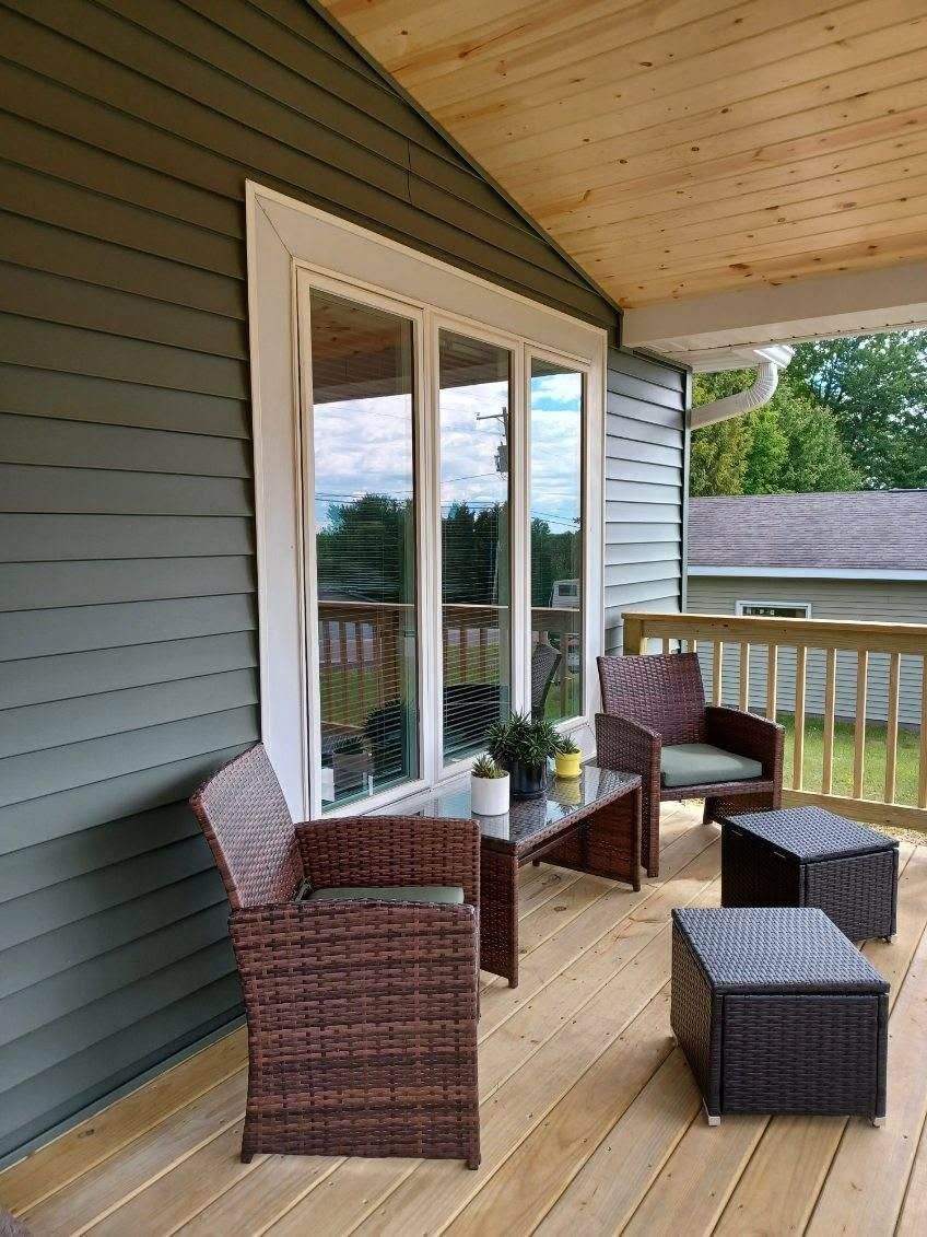 Covered porch with wicker chairs and ottomans beside gray siding and large windows.