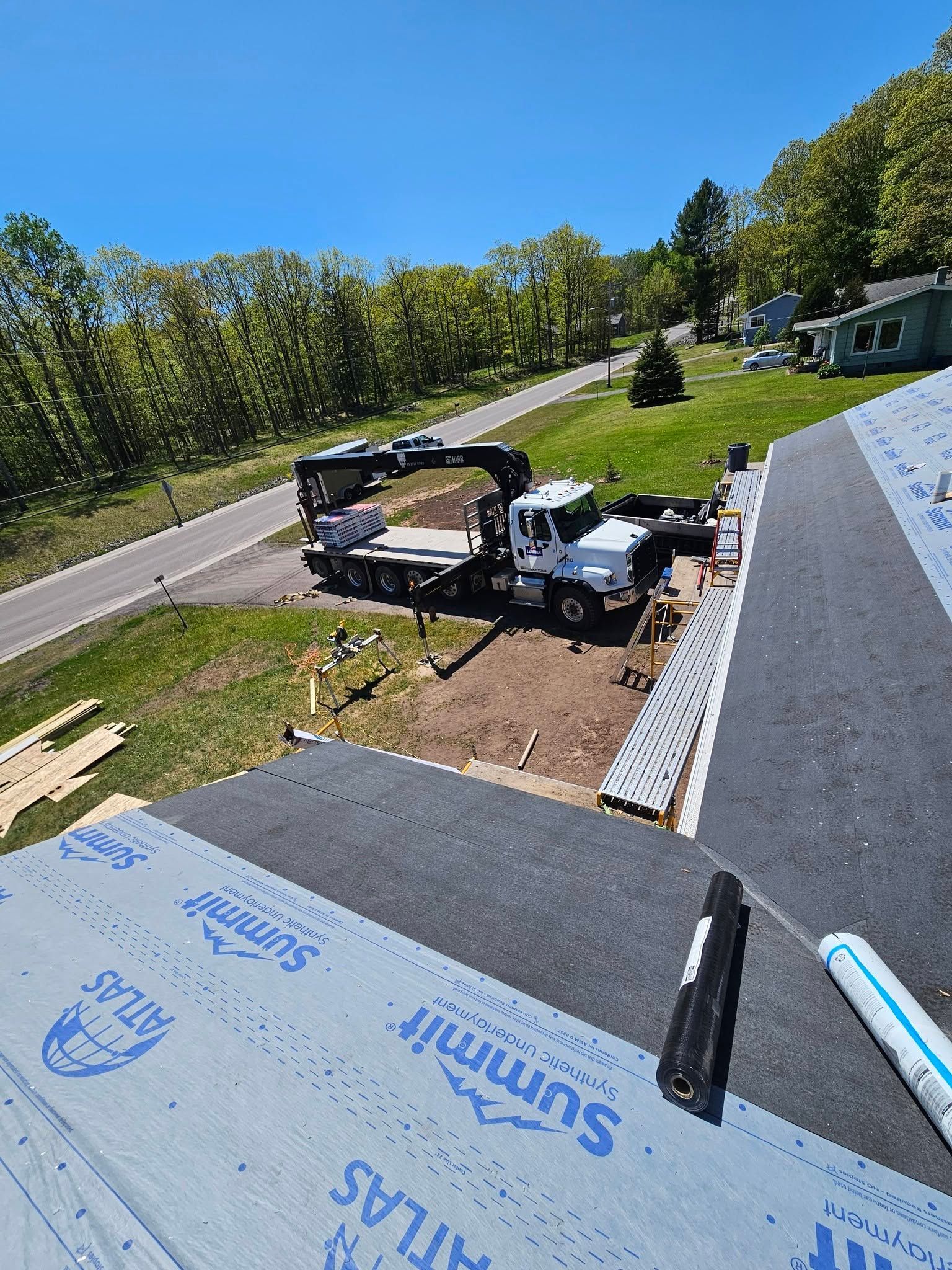 Roof under construction with workers, dark roofing underlayment, and a sloped driveway beside trees