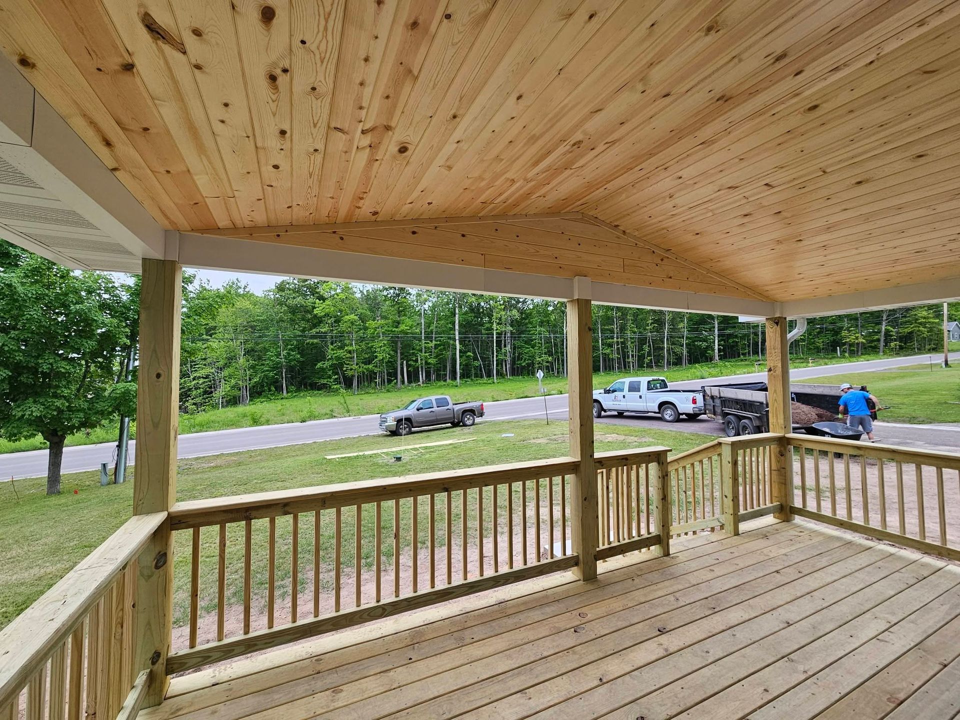 Wooden covered porch with railings overlooking a road and parked cars beside a tree-lined yard.