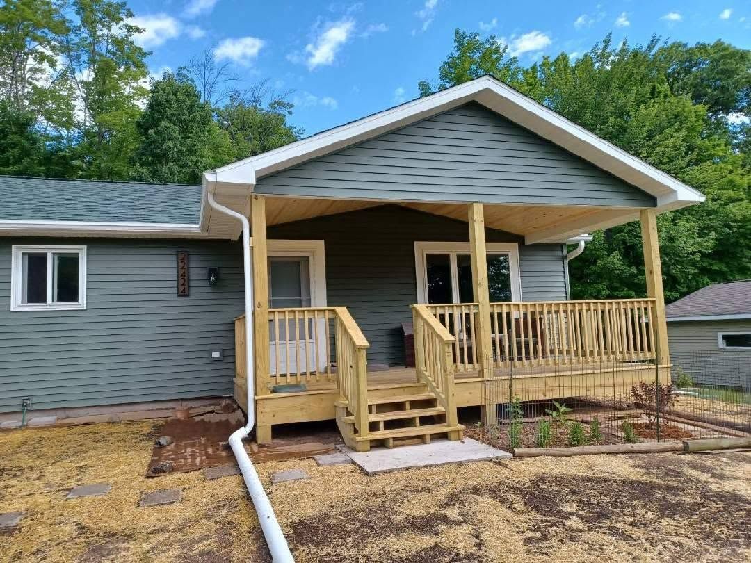 Front porch addition on a gray house, with wooden steps, railing, and a white roof under a blue sky.