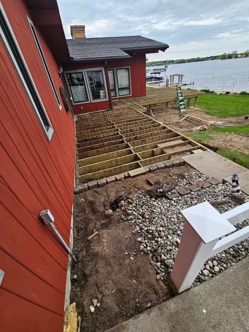Red lakeside house with a stepped dirt yard and a partially built wooden deck under construction