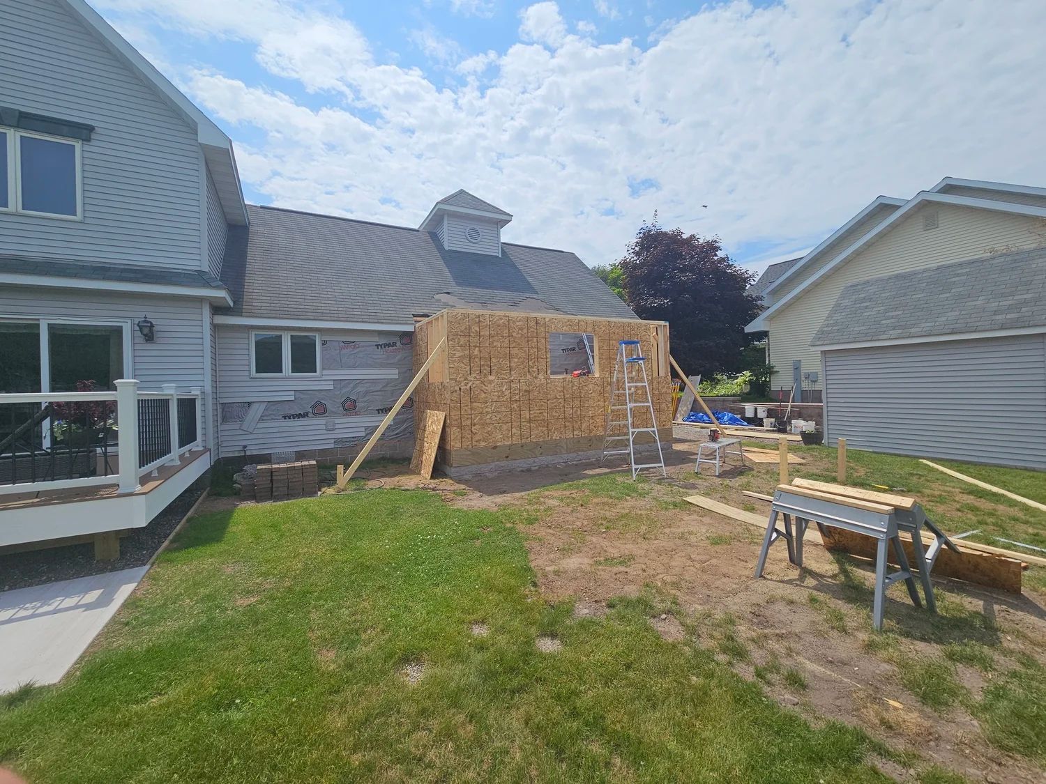 Backyard with a small unfinished wooden shed beside a house, lawn in foreground, and a workbench nearby
