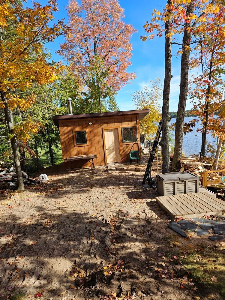 Small wooden cabin among autumn trees near a lake, with a sunny dirt yard and stacked pallets.