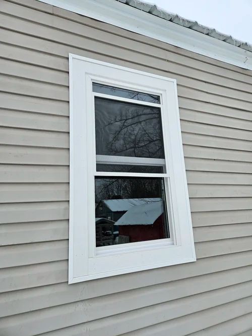 White-trimmed window on a beige house siding, reflecting trees and a snowy roof.