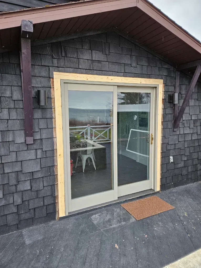 Sliding glass patio door on a shingled house with a roof overhang and deck railing visible through the glass.
