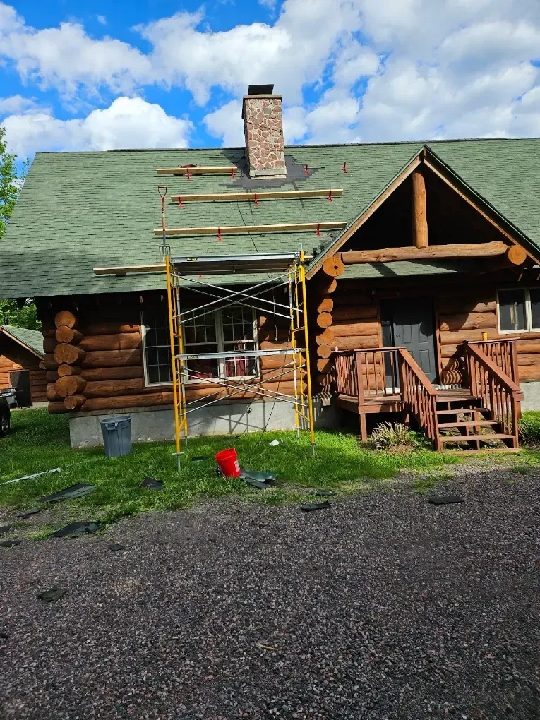 Log cabin with green roof and chimney under repair, scaffolding on the front porch, blue sky above