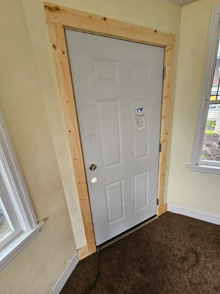 White door framed by unfinished wood in a carpeted room with yellow walls and a window.