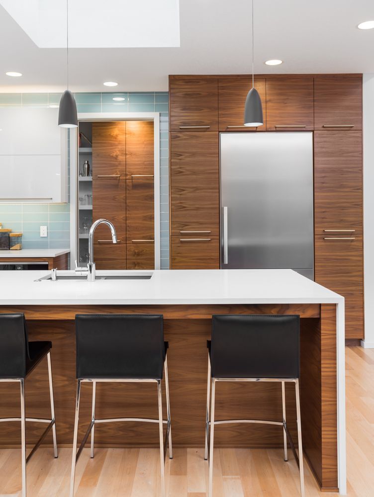 Modern kitchen with white island, three black stools, stainless-steel fridge, and wood-paneled cabinets.
