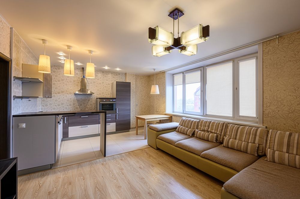Modern open-plan apartment with kitchen, brown sectional sofa, and large windows with balcony door.