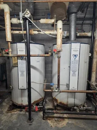 Two large white water heaters in a basement, with rusty pipes.