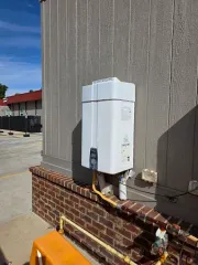 White outdoor water heater mounted on brick and siding exterior wall, connected to gas lines.