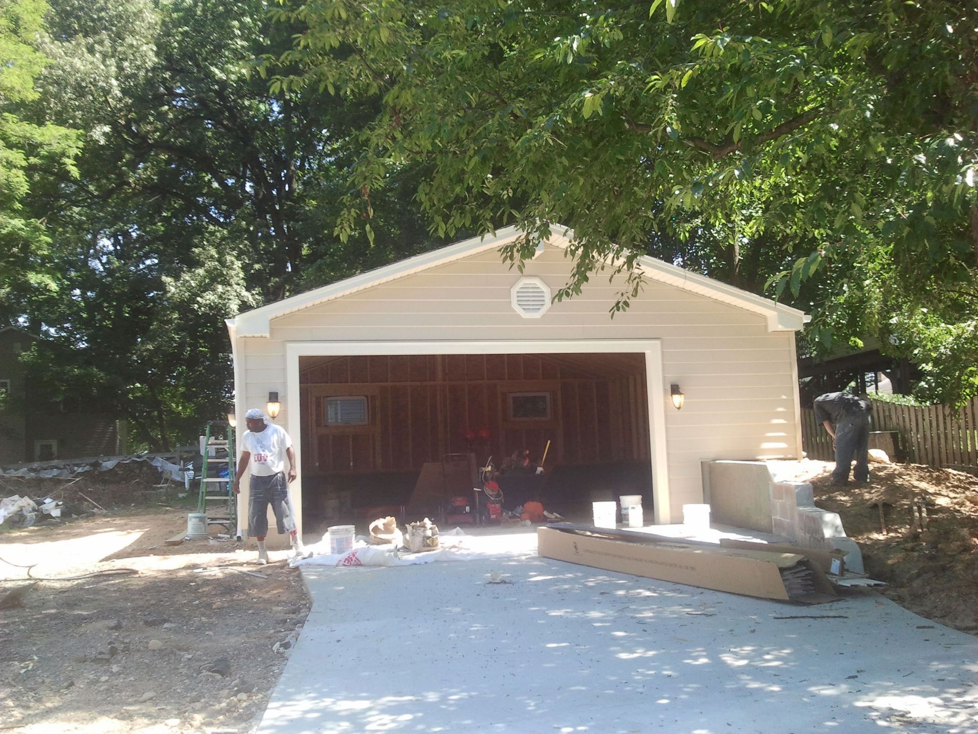 New garage construction; tan siding, open door, concrete driveway. Two people working outside.