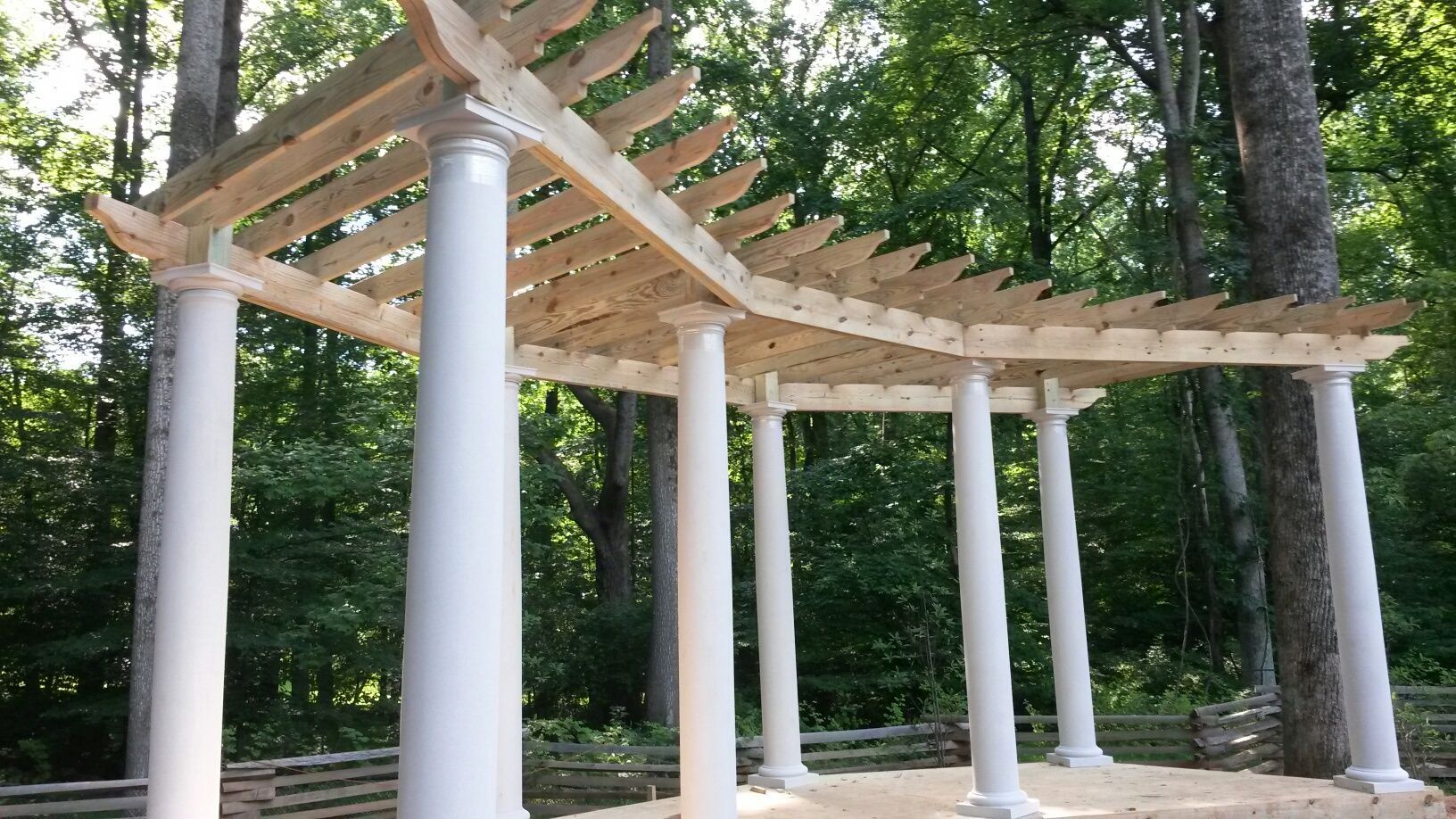 White pergola with wooden slats, supported by white columns, set in a wooded area.