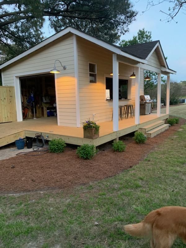 Yellow outbuilding with garage door and covered porch, on wooden deck. Green grass, brown mulch.