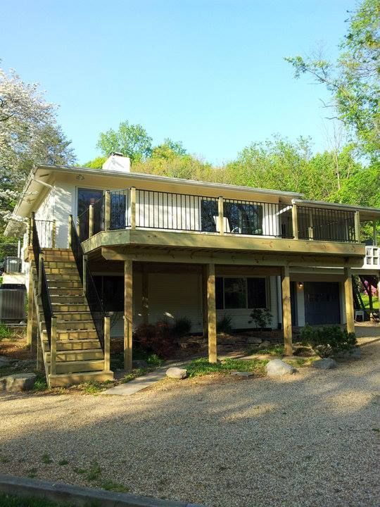 Two-story house with wooden decks and stairs, white siding, surrounded by trees, gravel driveway.