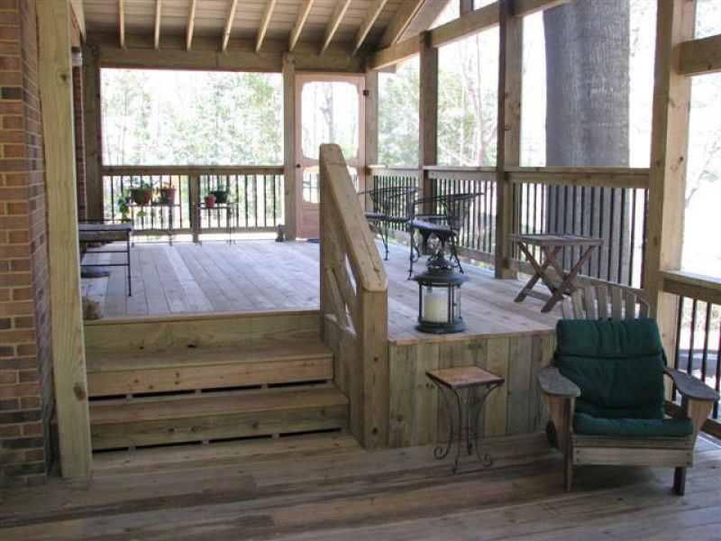 Wooden screened porch with stairs, railing, furniture, and a brick wall.
