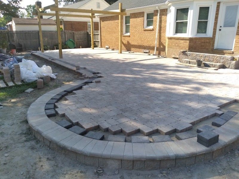 Construction of a patio with gray pavers and a curved border, near a brick house and wooden pergola.