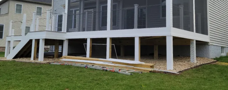A white screened porch on a house has a stairway and is surrounded by a gravel bed and grass.
