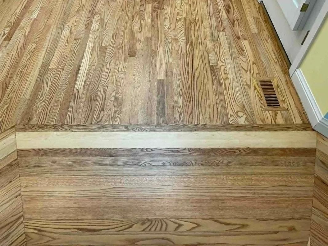 Wooden floor with a raised threshold between two rooms and a floor vent on the right.