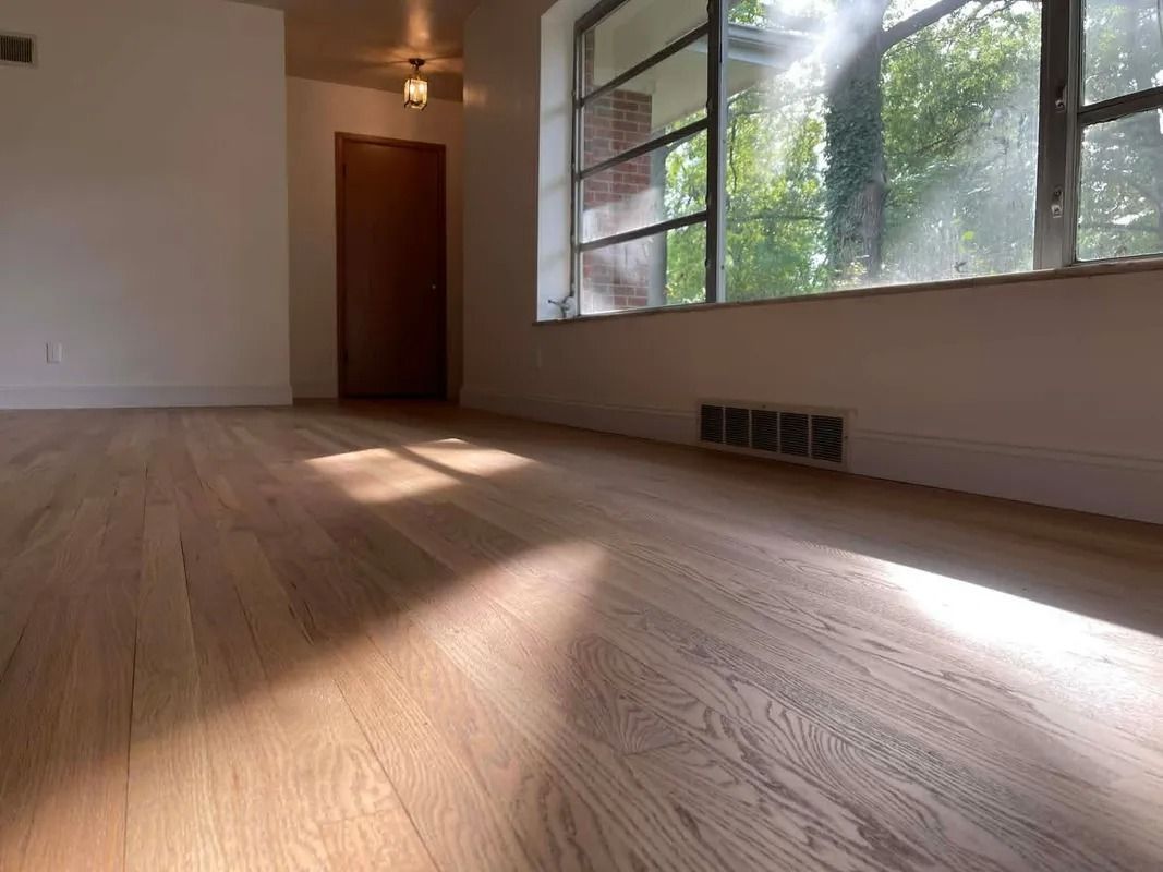 Empty sunlit room with wood floors, large windows, and a closed door.
