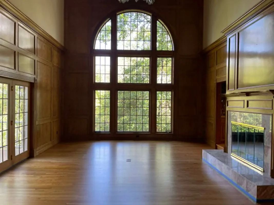 Empty sunlit room with hardwood floors, tall arched windows, and a mirrored wall fireplace