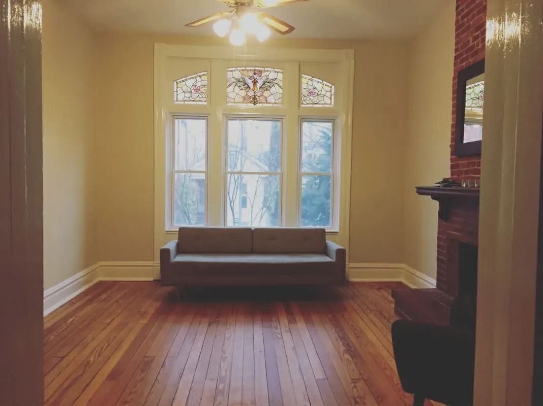 Empty living room with hardwood floors, large window seat, ceiling fan, and fireplace mantel