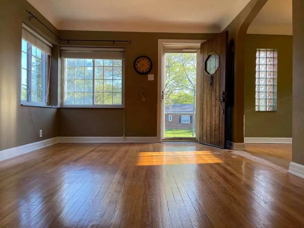 Empty living room with hardwood floors, tan walls, large windows, and sunlight on the floor