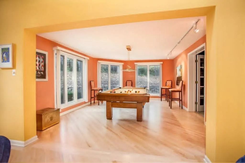 Bright game room with a pool table, orange walls, light wood floor, and large windows.
