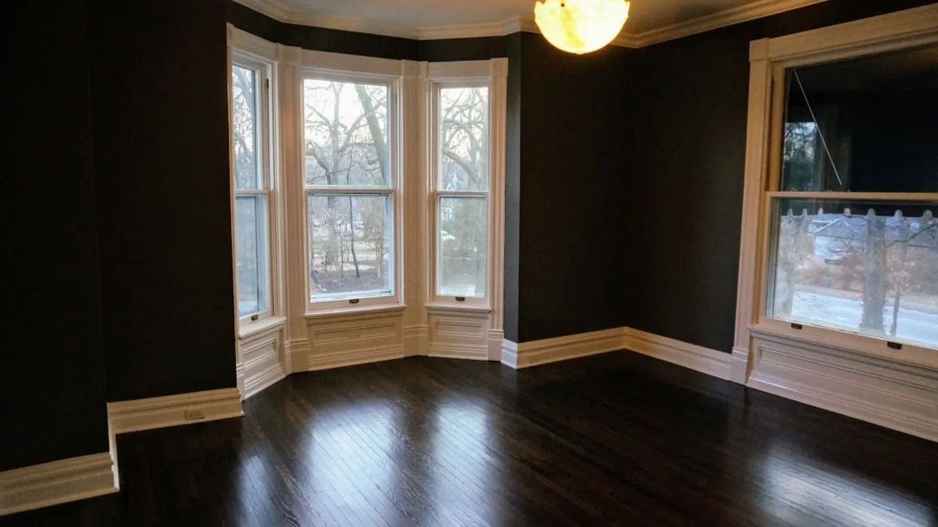 Empty room with dark hardwood floors, white trim, bay windows, and a hanging light fixture.