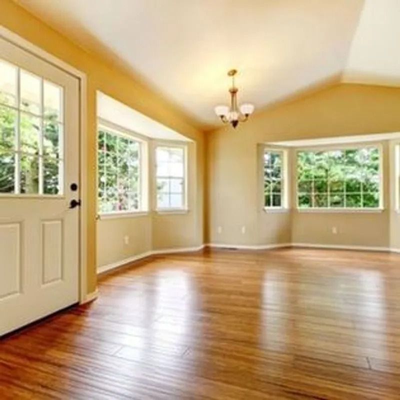 Empty sunlit room with hardwood floors, beige walls, and large windows beside a white door