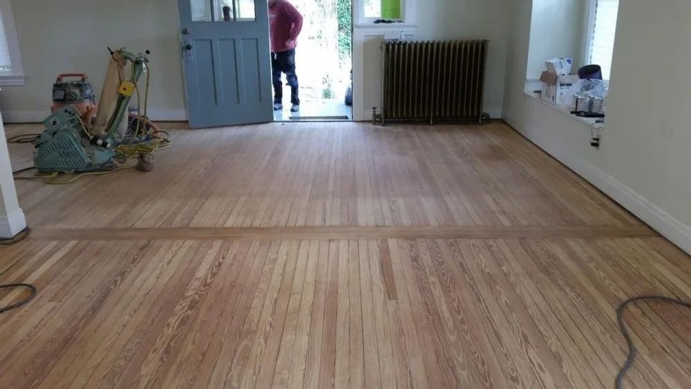 Room with newly laid wooden floorboards; person stands in doorway near a radiator and tools lie on the left.