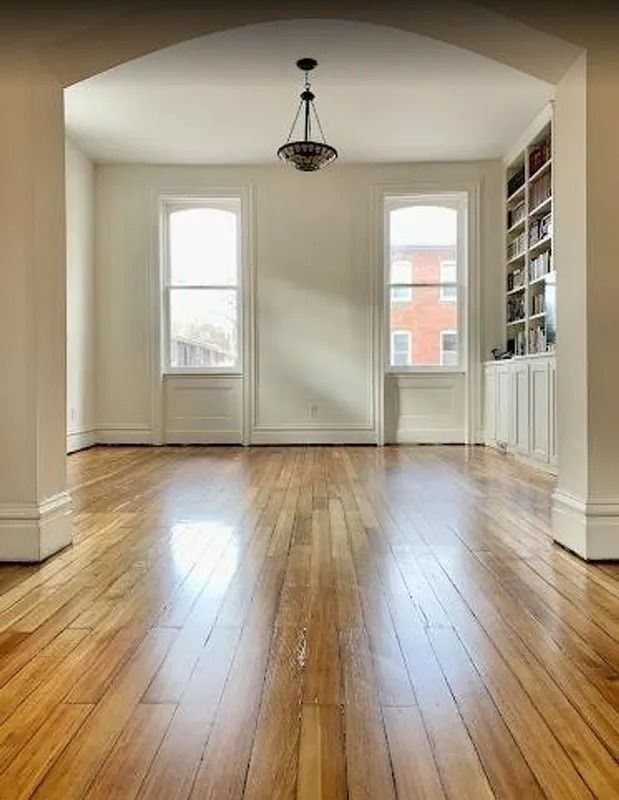 Empty room with polished wood floors, arched doorway, hanging light, and windows letting in daylight.