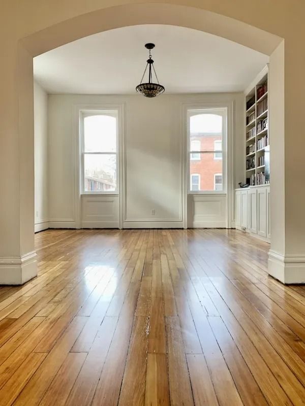 Empty bright room with hardwood floors, white walls, arched opening, and two tall windows.