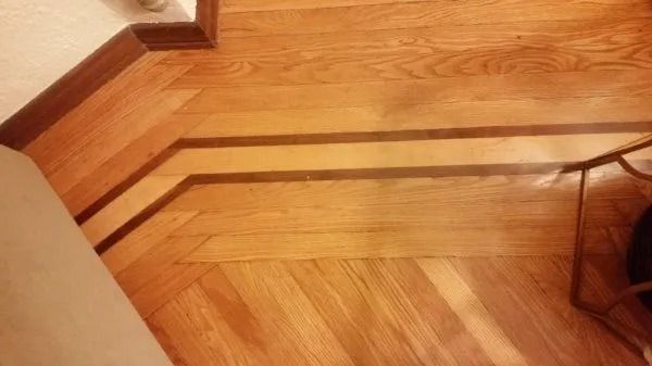 Wooden floor corner with a dark trim border and a black electrical cord along the wall.
