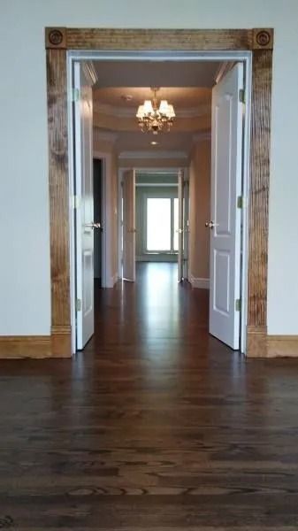 Elegant hallway with dark wood floors, white walls, ornate doorway, and chandelier leading to a bright room.