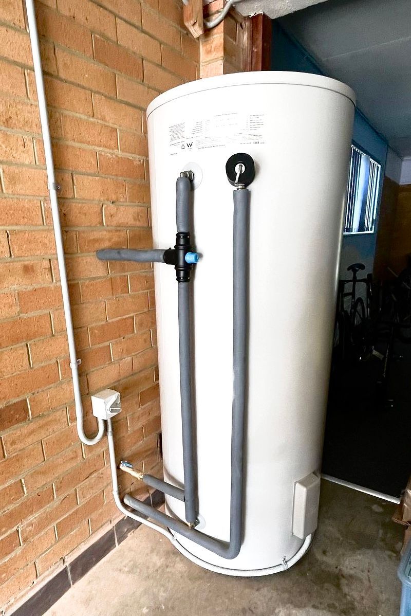 Large white cylindrical water heater beside a brick wall in a narrow utility area. — Hot Water Specialist Forster In Forster, NSW