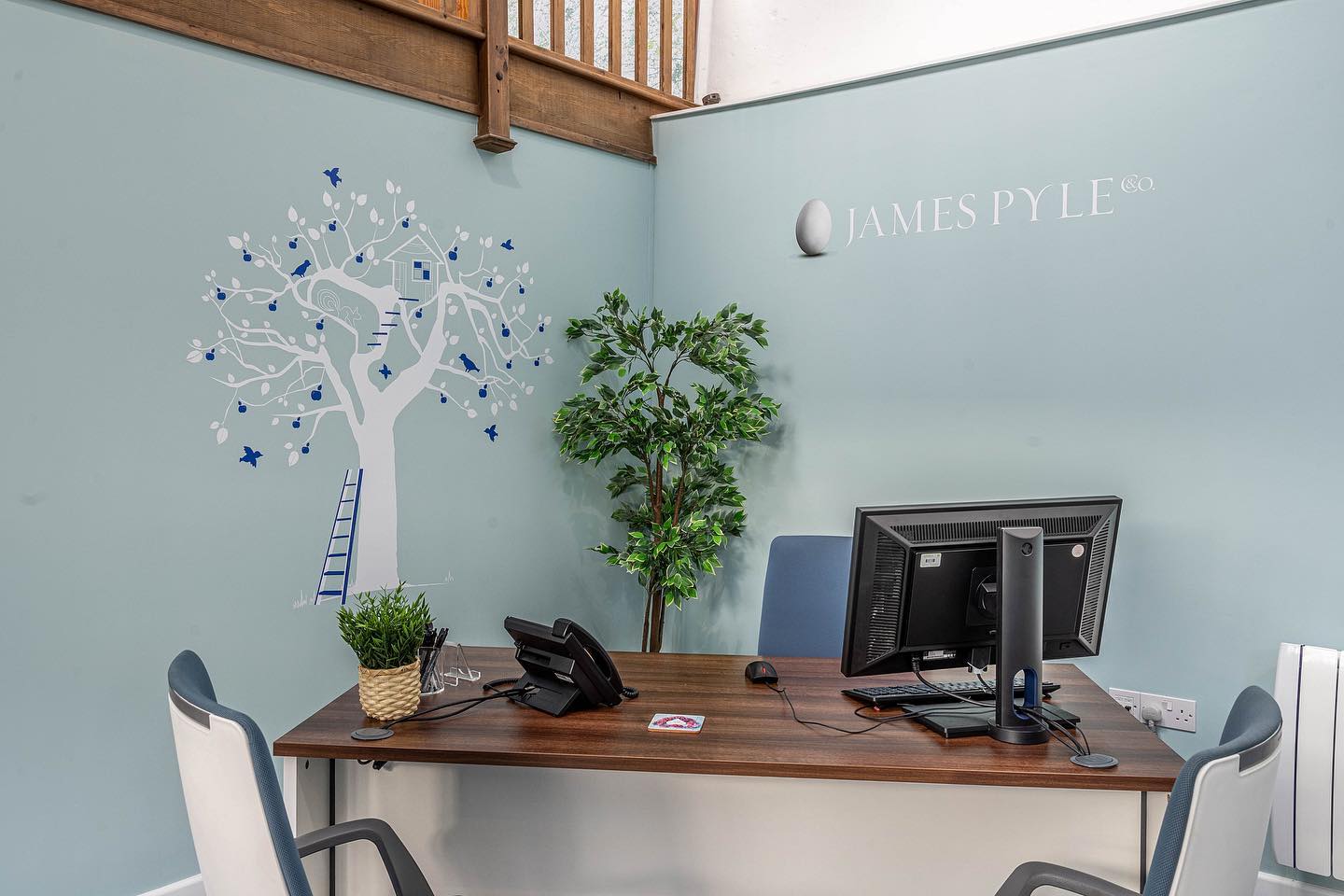 A desk with a computer and a phone on it in a room with a tree on the wall.