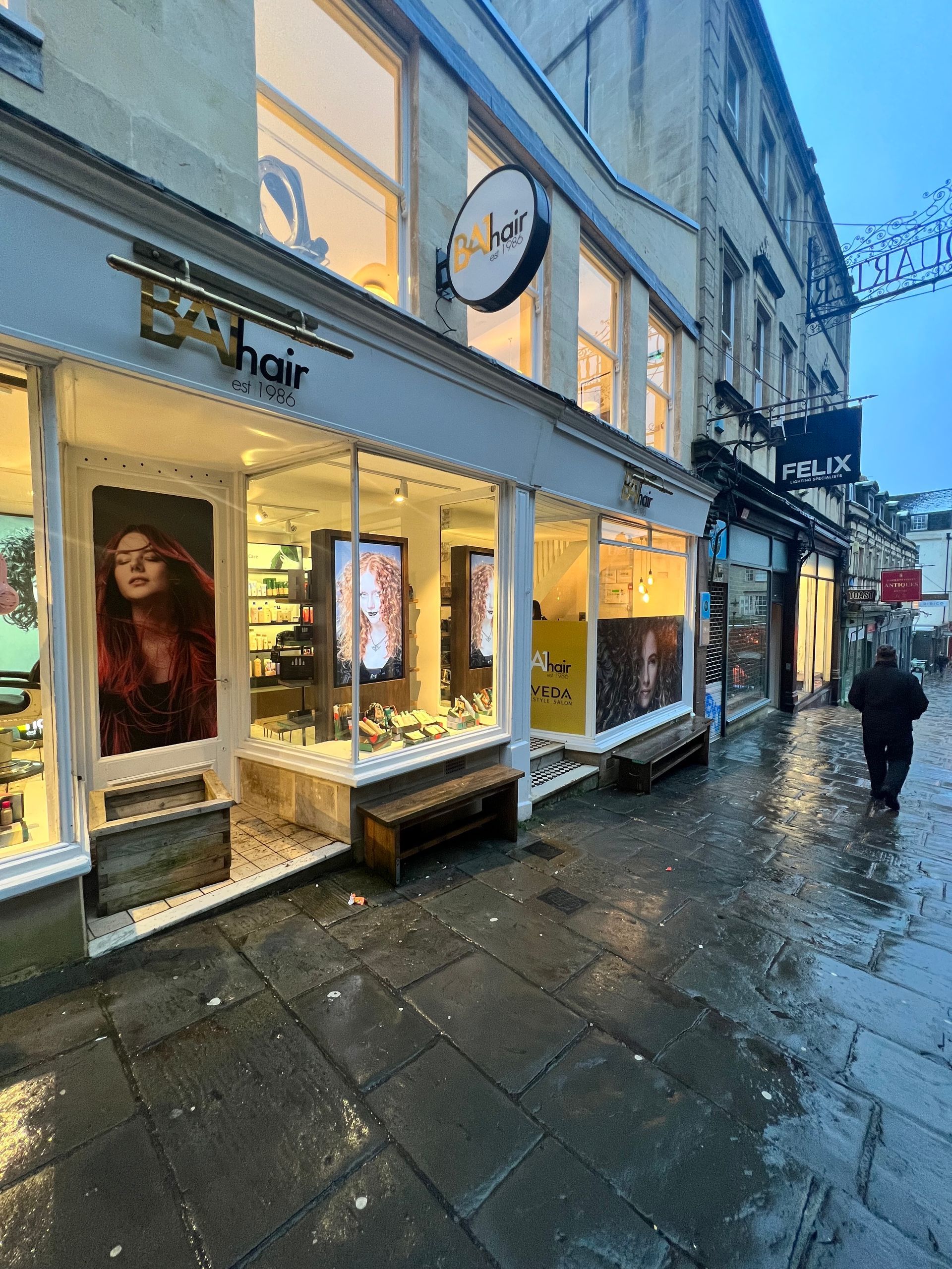 A man is walking down a wet sidewalk in front of a hair salon.