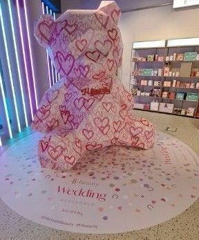 A pink teddy bear with hearts on it is sitting on a floor in a store.