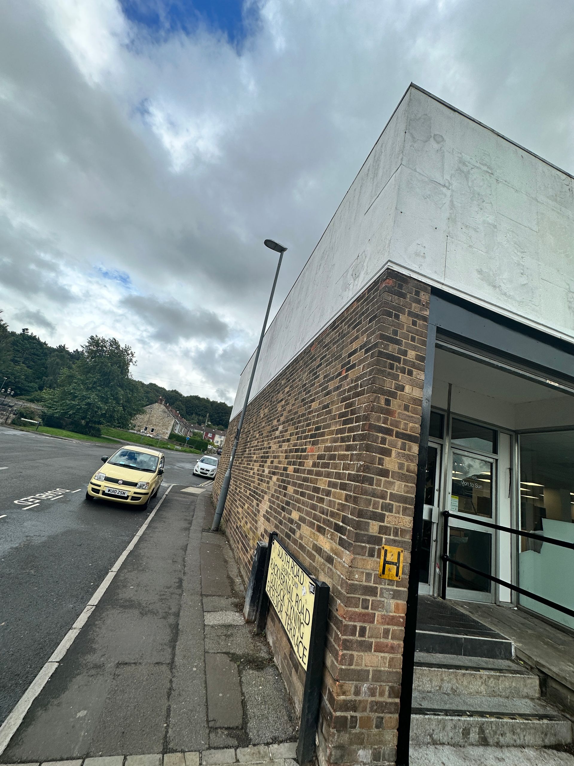 A yellow car is driving down a street next to a brick building.